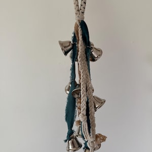 May include: A decorative hanging ornament featuring silver bells, teal and brown fabric strips, and a braided cream-colored rope. The bells are arranged in a cluster, creating a visually appealing and melodic accent.