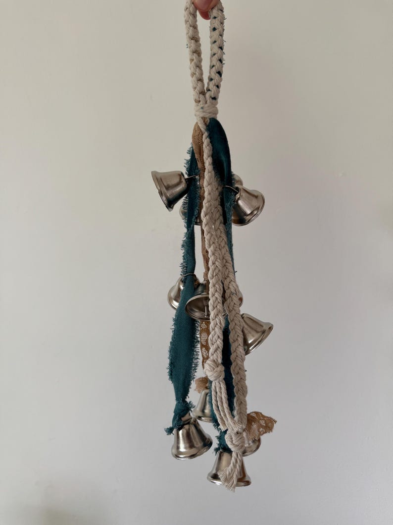 May include: A decorative hanging ornament featuring silver bells, teal and brown fabric strips, and a braided cream-colored rope. The bells are arranged in a cluster, creating a visually appealing and melodic accent.