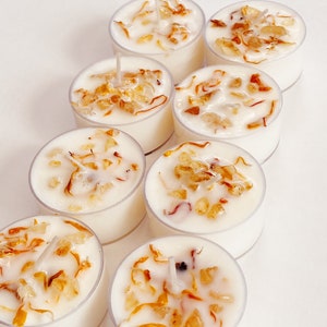 Peut inclure: Huit bougies chauffe-plat blanches avec une cire transparente et des fleurs s&eacute;ch&eacute;es et des cristaux incrust&eacute;s. Les bougies sont dispos&eacute;es en rang&eacute;e sur une surface blanche.