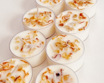 May include: Eight white tea light candles with a clear wax and embedded dried flowers and crystals. The candles are arranged in a row on a white surface.