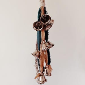 May include: A decorative hanging ornament featuring several silver bells suspended from a macrame cord and fabric strips in shades of teal, beige, and brown. The bells are clustered together, creating a visually appealing and textured design.