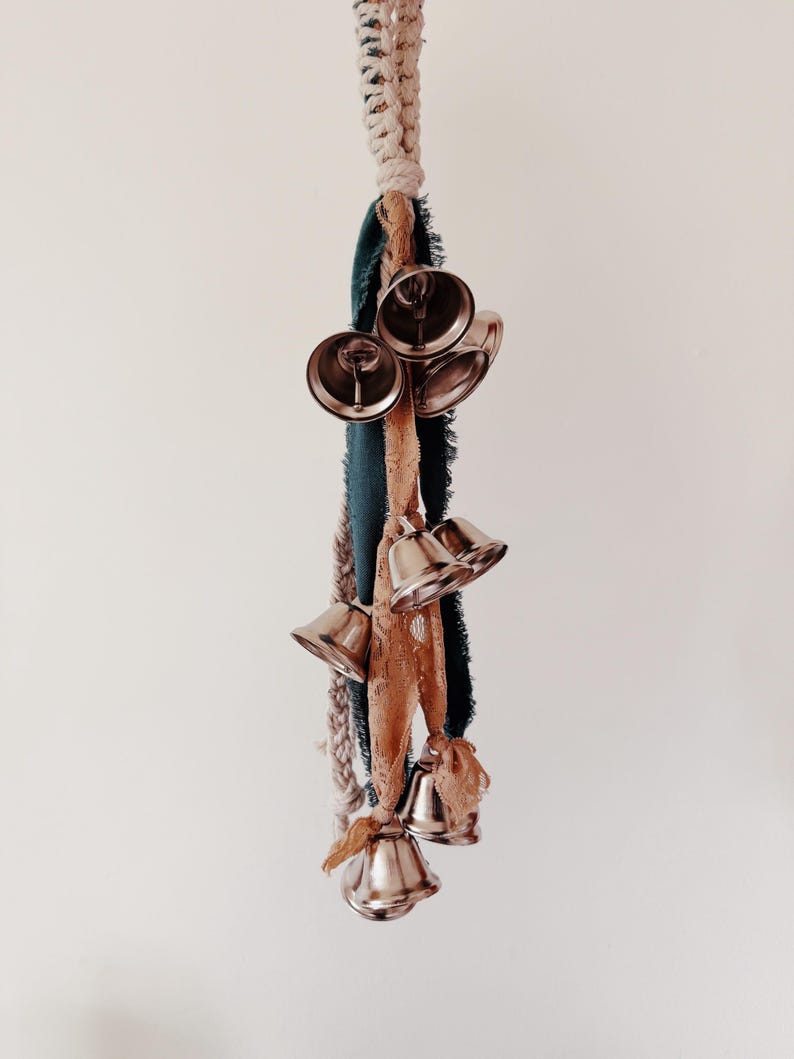 May include: A decorative hanging ornament featuring several silver bells suspended from a macrame cord and fabric strips in shades of teal, beige, and brown. The bells are clustered together, creating a visually appealing and textured design.