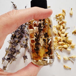 May include: A clear glass bottle with a black dropper top, filled with a golden liquid and botanical inclusions. The bottle is held by a hand, with sprigs of lavender and dried jasmine flowers in the background. The bottle's contents appear to be a beauty product.