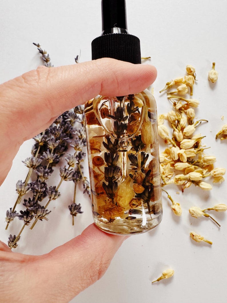May include: A clear glass bottle with a black dropper top, filled with a golden liquid and botanical inclusions. The bottle is held by a hand, with sprigs of lavender and dried jasmine flowers in the background. The bottle's contents appear to be a beauty product.