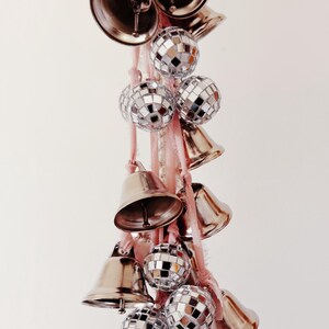 May include: A string of silver bells and disco ball ornaments hanging from a pink ribbon. The bells are various sizes and shapes, and the disco balls are all the same size.