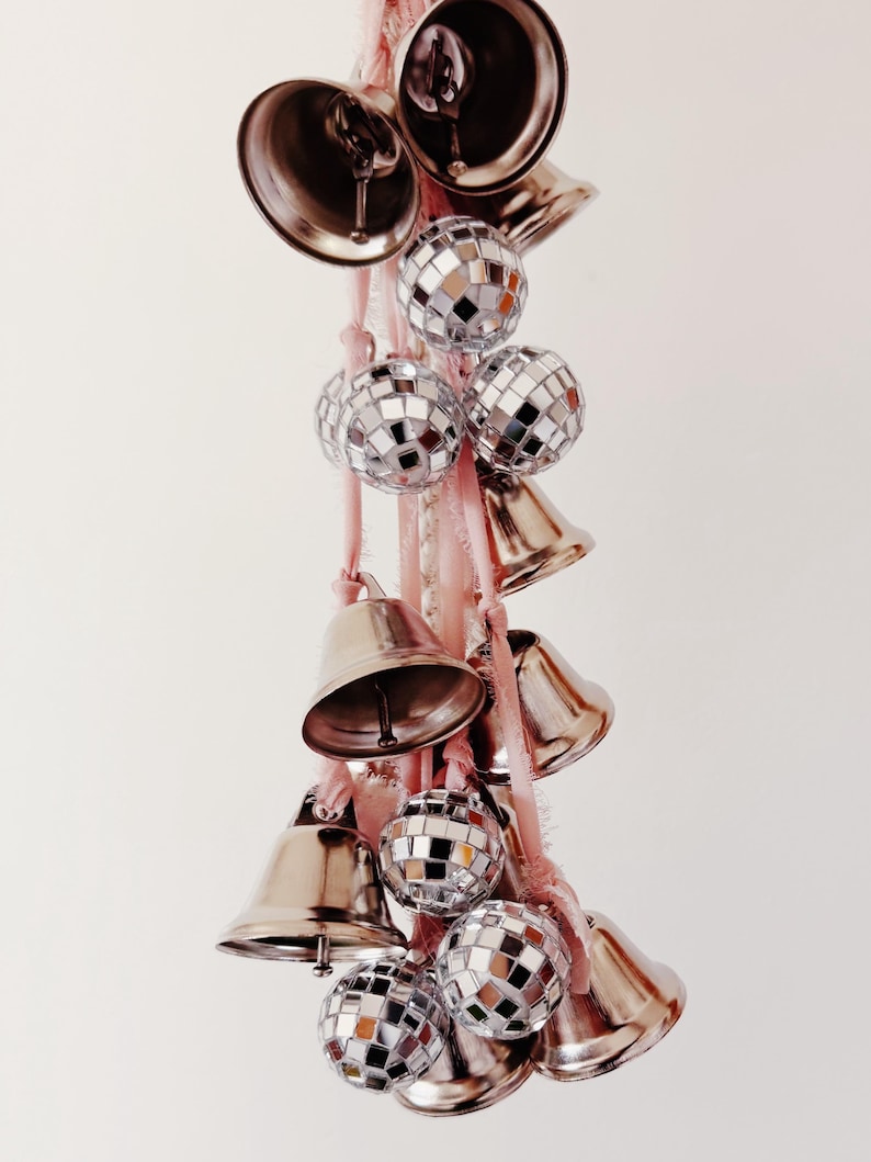 May include: A string of silver bells and disco ball ornaments hanging from a pink ribbon. The bells are various sizes and shapes, and the disco balls are all the same size.