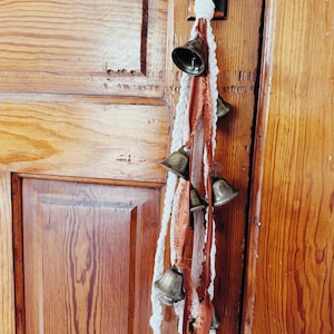 May include: A white rope with brown and white fabric tassels hangs from a wooden door. The rope has a series of small, gold-colored bells attached to it.