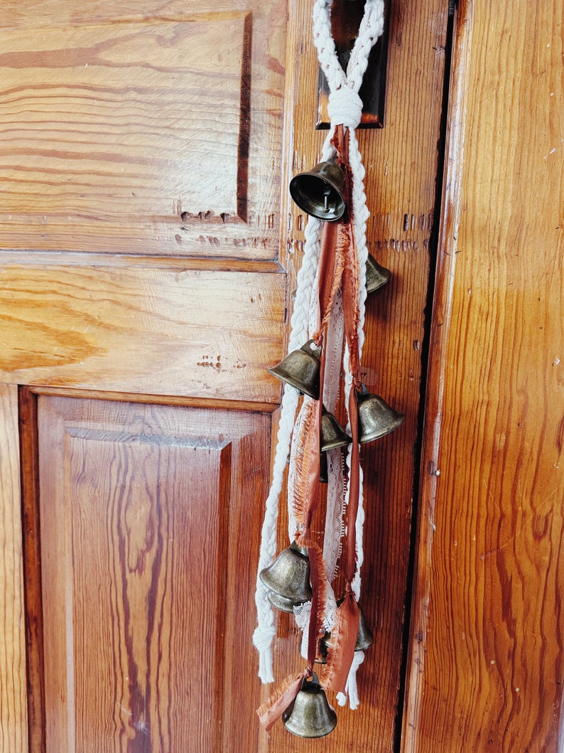 May include: A white rope with brown and white fabric tassels hangs from a wooden door. The rope has a series of small, gold-colored bells attached to it.