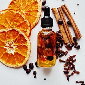 May include: A clear glass bottle with a black dropper, filled with a golden liquid and botanicals, sits beside dried orange slices, coffee beans, cinnamon sticks, and cloves. The ingredients are arranged on a white surface.