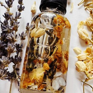 May include: A clear glass bottle with a black dropper top filled with dried flowers and herbs. The bottle is surrounded by sprigs of lavender and other dried flowers, creating a natural and botanical aesthetic.