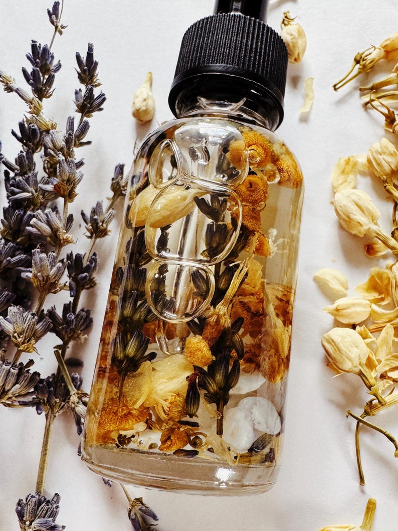 May include: A clear glass bottle with a black dropper top filled with dried flowers and herbs. The bottle is surrounded by sprigs of lavender and other dried flowers, creating a natural and botanical aesthetic.