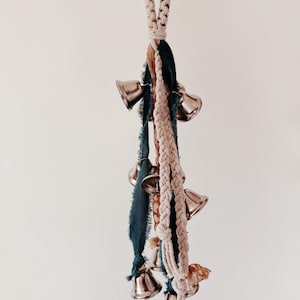 May include: A decorative hanging ornament featuring multiple silver bells. The bells are suspended from a braided rope and accented with teal and tan fabric strips. The ornament is designed for home decor.