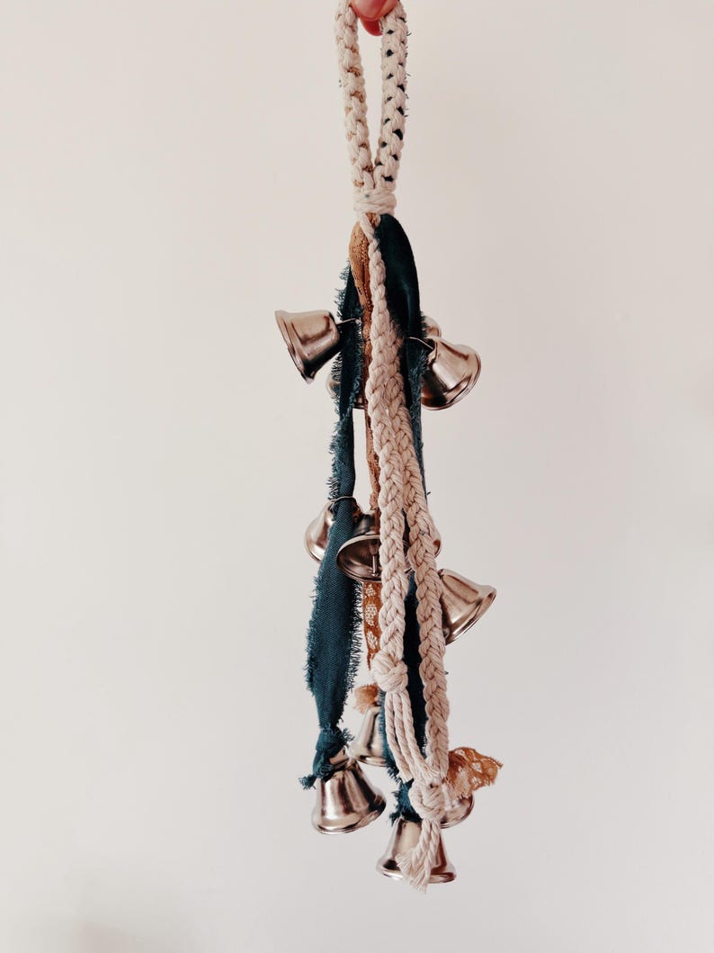 May include: A decorative hanging ornament featuring multiple silver bells. The bells are suspended from a braided rope and accented with teal and tan fabric strips. The ornament is designed for home decor.
