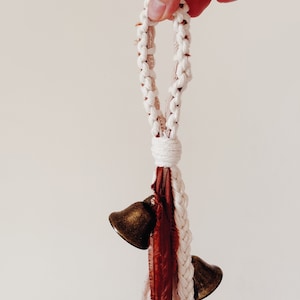 May include: A white macrame rope with a loop at the top and two small brass bells hanging from the bottom. The rope is also decorated with a long, thin piece of rust-colored fabric.