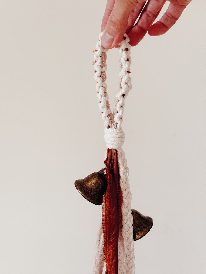 May include: A white macrame rope with a loop at the top and two small brass bells hanging from the bottom. The rope is also decorated with a long, thin piece of rust-colored fabric.