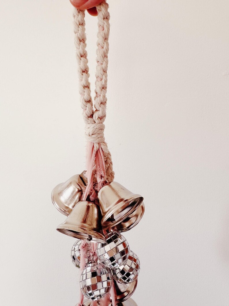 May include: A white rope with a pink tassel hangs from the ceiling. Attached to the rope are a cluster of silver bells and disco ball ornaments.