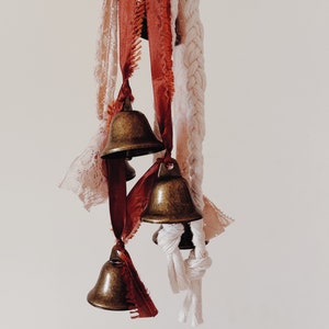 May include: A collection of small brass bells hanging from a white rope and red and white ribbons. The bells are tied together with knots and the ribbons are frayed at the ends.