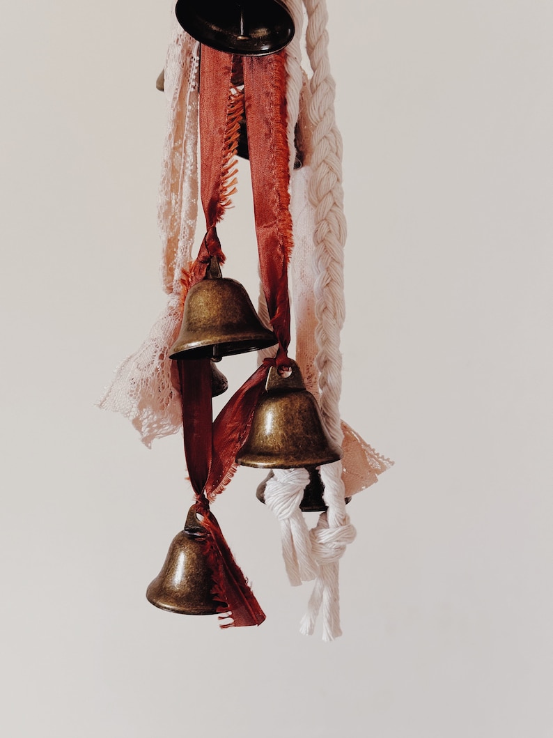 May include: A collection of small brass bells hanging from a white rope and red and white ribbons. The bells are tied together with knots and the ribbons are frayed at the ends.