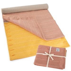 May include: A rolled-up yoga mat with a light tan exterior, and a pink and yellow yoga mat with white stripes and tassels. A folded pink yoga mat is tied with a cream ribbon.