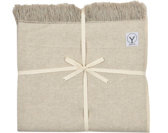 May include: A folded, beige-colored textile with fringed edges, tied with a cream-colored ribbon. A small white tag with the word "GAYO" is visible. The textile appears to be a blanket or throw.