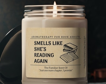 May include: A lit candle in a glass jar with a lid. The label reads "Smells Like She's Reading Again" with the text "Aromatherapy for Book Addicts." The candle has a neutral color and a simple design, perfect for book lovers.