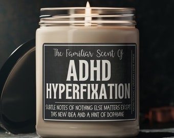 May include: A white candle with a black label that reads "The Familiar Scent Of ADHD HYPERFIXATION. Subtle notes of nothing else matters except this new idea and a hint of dopamine."