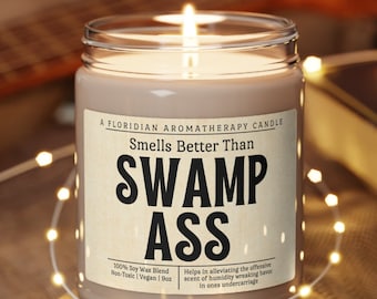 May include: A brown candle with a white label that reads "A Floridian Aromatherapy Candle Smells Better Than Swamp Ass 100% Soy Wax Blend Non-Toxic | Vegan | 8oz Helps in alleviating the offensive scent of humidity wreaking havoc in ones undercarriage."