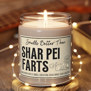 May include: A brown candle with a white label that reads "Smells Better Than Shar Pei Farts" with a cartoon dog and a poop emoji. The candle is on a wooden coaster.