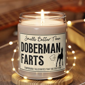 May include: A brown glass candle with a white label that reads "Smells Better Than Doberman Farts Temporarily Alleviates That Big Dog Stink". The candle is lit and sitting on a wooden coaster.