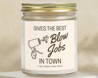 May include: A clear glass candle jar with a gold lid. The label reads "Gives the best Blow Jobs in Town" with a hairdryer graphic. The candle is 9 oz, vegan, and non-toxic.