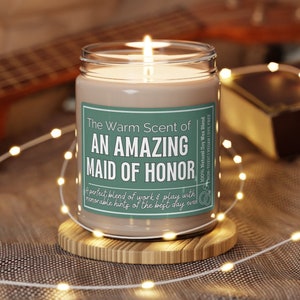 May include: A green and white candle jar with the text "The Warm Scent of An Amazing Maid of Honor" and "A perfect blend of work & play with memorable hints of the best day ever". The candle is on a wooden coaster and surrounded by white fairy lights.
