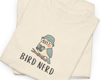 May include: Two cream-colored t-shirts are stacked. The top shirt features a cartoon bird wearing glasses and a hat, holding binoculars, perched on a branch. The text "BIRD NERD" is below the bird in black letters.