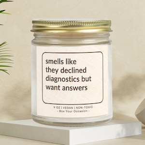 May include: A glass candle jar with a gold lid. The label reads "smells like they declined diagnostics but want answers" and "9 OZ | VEGAN | NON-TOXIC - Box Your Occasion - "