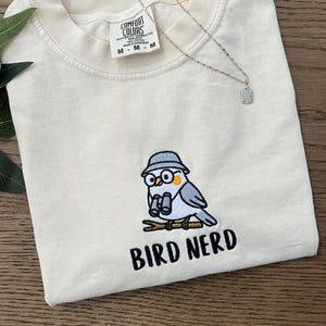 May include: Cream-colored t-shirt with an embroidered cartoon bird wearing a hat and glasses, holding binoculars. The text "BIRD NERD" is below the bird. A silver necklace with a pendant is also visible.