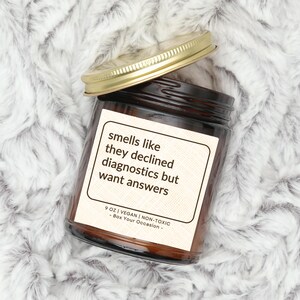 May include: A brown glass candle jar with a gold lid. The label reads "smells like they declined diagnostics but want answers" and "9 oz | Vegan | Non-Toxic - Box Your Occasion - "