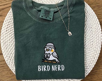 May include: A dark green t-shirt with a cartoon bird design and the text "BIRD NERD" in white. The bird is wearing a hat and glasses. A silver necklace with a small pendant is also present.