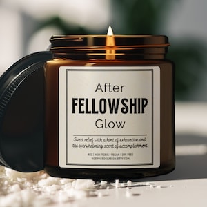 May include: A brown glass candle jar with a white label that reads "After Fellowship Glow". The label also includes the text "Sweet relief with a hint of exhaustion and the overwhelming scent of accomplishment". The candle is lit and the flame is visible.