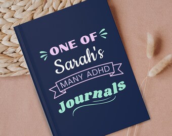 May include: A dark blue journal with the text "ONE OF Sarah's MANY ADHD Journals" in pink and light blue lettering.