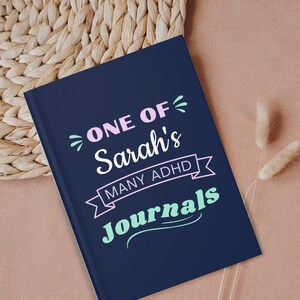 May include: A dark blue journal with the text "ONE OF Sarah's MANY ADHD Journals" in pink and light blue lettering.