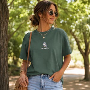 May include: A dark green t-shirt with the embroidered text "BIRD NERD" below a small cartoon bird wearing a hat. The shirt is paired with blue jeans. The image is taken outdoors with a blurred green background.