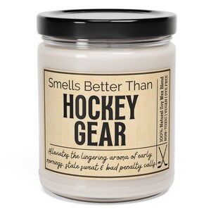 May include: A glass jar candle with a brown label that reads "Smells Better Than HOCKEY GEAR Alleviates the lingering aroma of early mornings. stale sweat & bad penalty calls!"  The label also includes the text "100% Natural Soy Wax Blend NON-TOXIC | VEGAN | DYE FREE".
