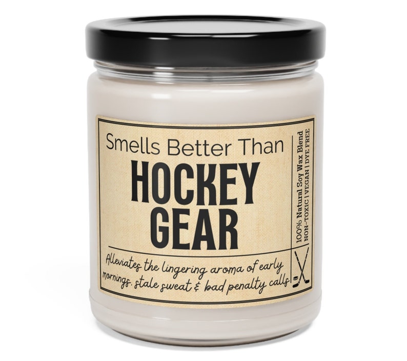 May include: A glass jar candle with a brown label that reads "Smells Better Than HOCKEY GEAR Alleviates the lingering aroma of early mornings. stale sweat & bad penalty calls!"  The label also includes the text "100% Natural Soy Wax Blend NON-TOXIC | VEGAN | DYE FREE".