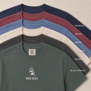 May include: A collection of Comfort Colors t-shirts in various colors, including midnight blue, brick red, denim blue, natural, pepper, and blue spruce. The bottom shirt features an embroidered bird with glasses and the text "BIRD NERD".