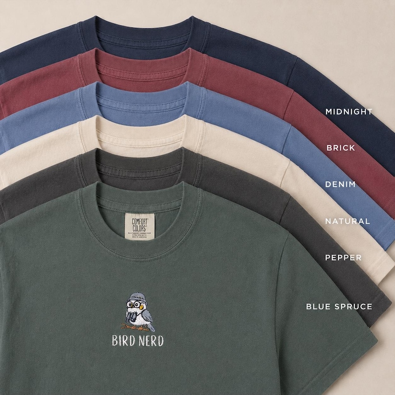 May include: A collection of Comfort Colors t-shirts in various colors, including midnight blue, brick red, denim blue, natural, pepper, and blue spruce. The bottom shirt features an embroidered bird with glasses and the text "BIRD NERD".
