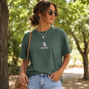 May include: A dark green t-shirt with the embroidered text "BIRD NERD" below a small cartoon bird wearing a hat. The shirt is paired with blue jeans. The image is taken outdoors with a blurred green background.