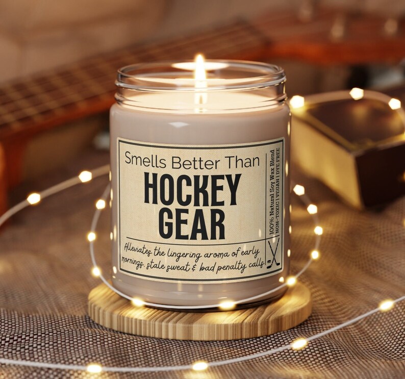 May include: A brown soy wax candle with a white label that reads "Smells Better Than Hockey Gear" and "Alleviates the lingering aroma of early mornings, stale sweat & bad penalty calls". The candle is sitting on a wooden coaster.
