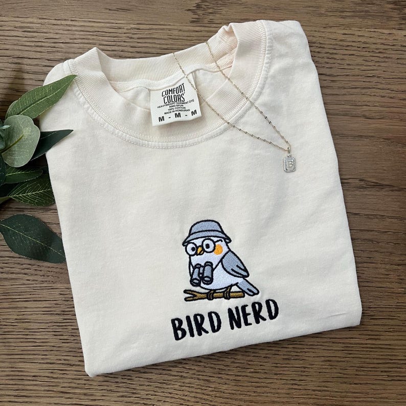 May include: Cream-colored Comfort Colors t-shirt with an embroidered bird design. The bird wears a hat and glasses, holding binoculars. The text "BIRD NERD" is below the bird. A silver necklace with a pendant is also visible.