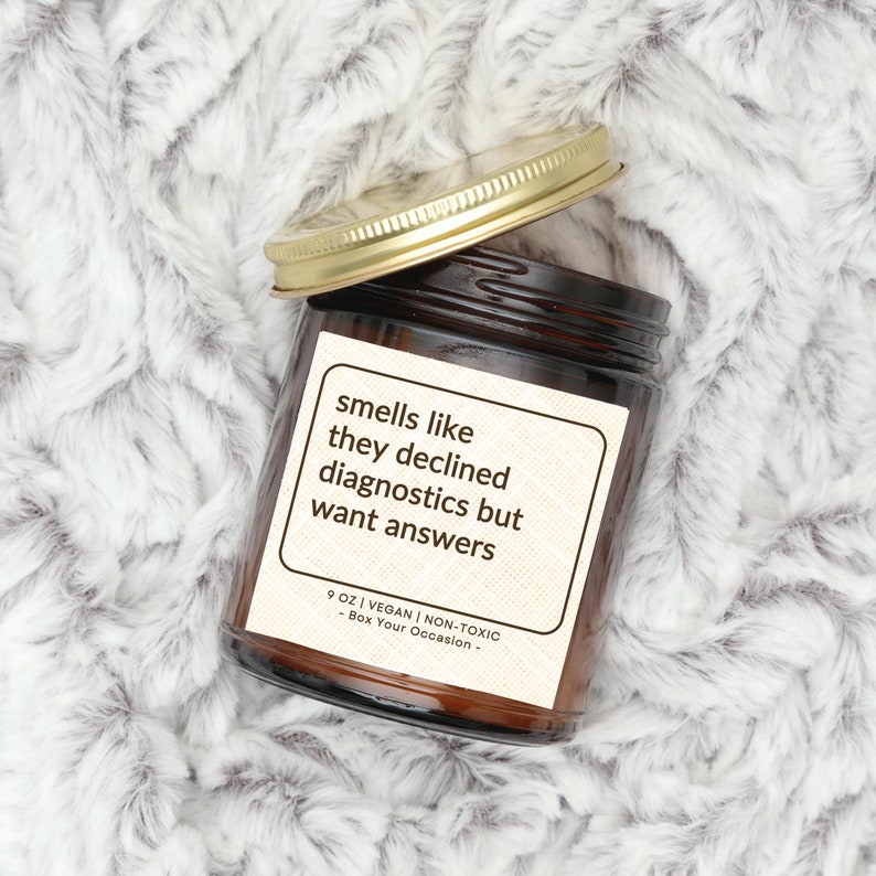 May include: A brown glass candle jar with a gold lid. The label reads "smells like they declined diagnostics but want answers" and "9 oz | Vegan | Non-Toxic - Box Your Occasion - "