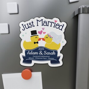 K&ouml;nnte beinhalten: Ein wei&szlig;er Magnet mit einem blauen Band und dem Text "Just Married" in Blau. Zwei gelbe Gummienenten mit schwarzen Augen und Schn&auml;beln k&uuml;ssen sich. Die m&auml;nnliche Ente tr&auml;gt einen schwarzen Zylinder und eine Fliege. Die weibliche Ente tr&auml;gt einen wei&szlig;en Schleier und eine rosafarbene Blumenkrone. Der Text "2024 Adam & Sarah Carnival Celebration Miami - Princess Cays - Nassau" steht auf dem Band.