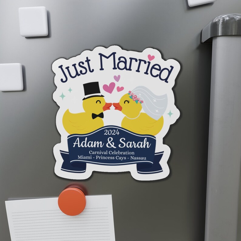 K&ouml;nnte beinhalten: Ein wei&szlig;er Magnet mit einem blauen Band und dem Text "Just Married" in Blau. Zwei gelbe Gummienenten mit schwarzen Augen und Schn&auml;beln k&uuml;ssen sich. Die m&auml;nnliche Ente tr&auml;gt einen schwarzen Zylinder und eine Fliege. Die weibliche Ente tr&auml;gt einen wei&szlig;en Schleier und eine rosafarbene Blumenkrone. Der Text "2024 Adam & Sarah Carnival Celebration Miami - Princess Cays - Nassau" steht auf dem Band.